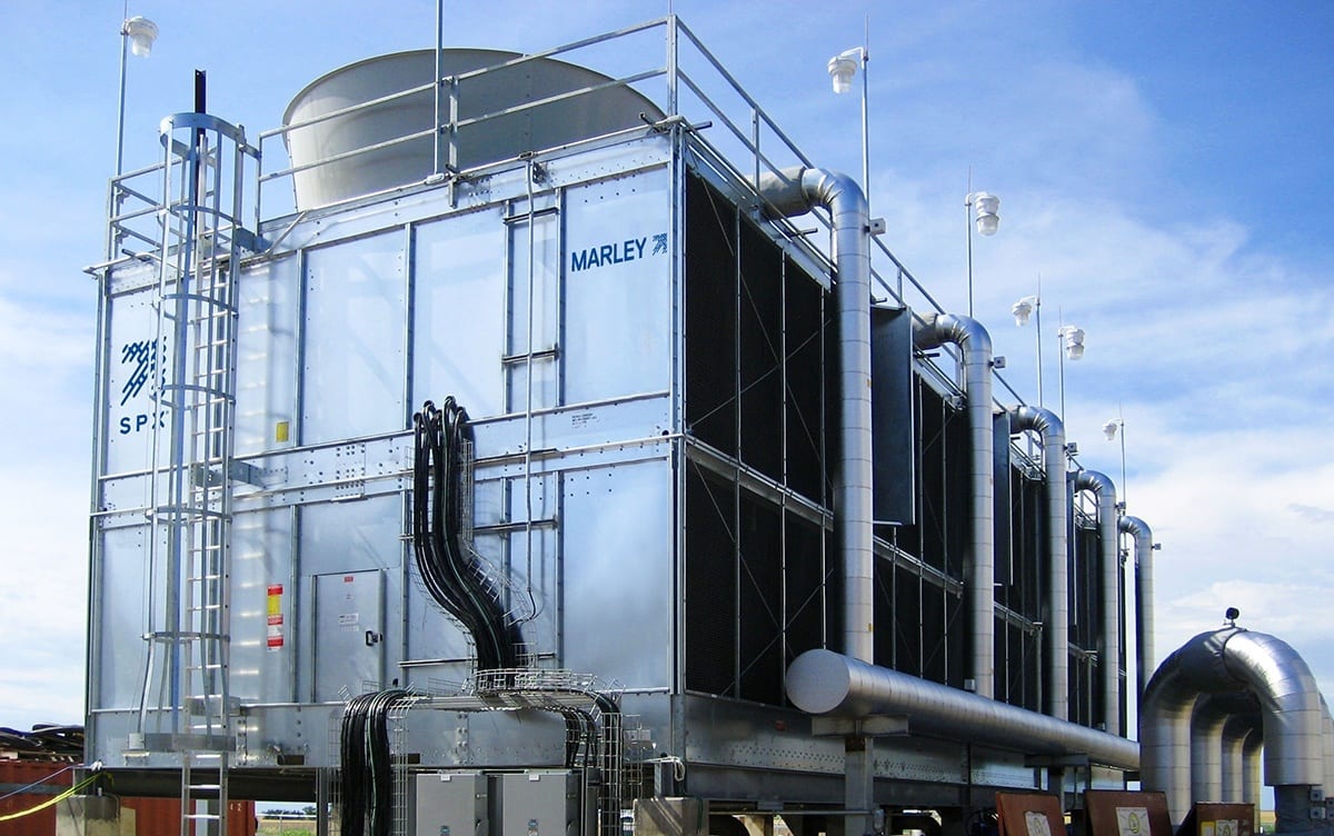 Large industrial cooling tower outdoors with metal structure, pipes, and a cloudy sky background.