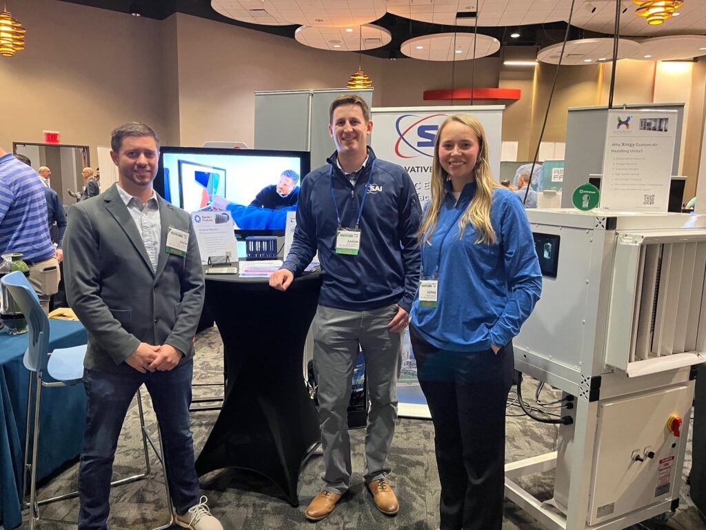 Three people standing in front of a technology booth at a conference. A male in a dark jacket and jeans, another male in a branded pullover, and a female in a similar pullover are posing for the picture. A monitor displaying a video and some informational signage are visible on the booth behind them. The setup is in a room with a patterned carpet and hanging ceiling lights.