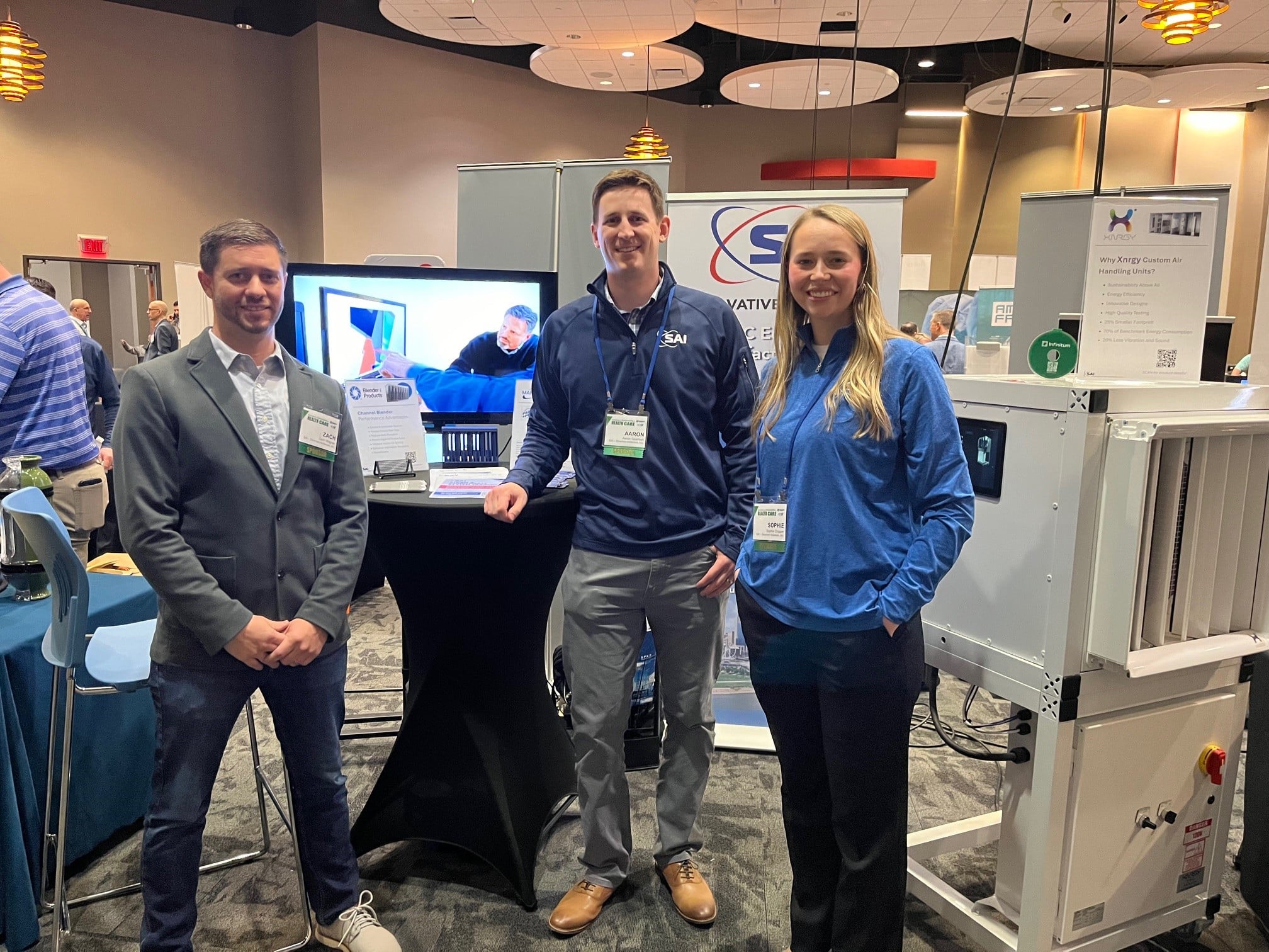 Three people standing in front of a technology booth at a conference. A male in a dark jacket and jeans, another male in a branded pullover, and a female in a similar pullover are posing for the picture. A monitor displaying a video and some informational signage are visible on the booth behind them. The setup is in a room with a patterned carpet and hanging ceiling lights.