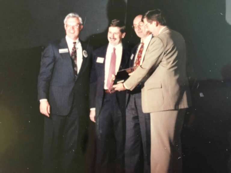 Four men in suits gathered on stage during a business award ceremony. Three of them are holding a trophy while smiling. They appear to be in a celebratory mood as they stand close together in a spotlight.