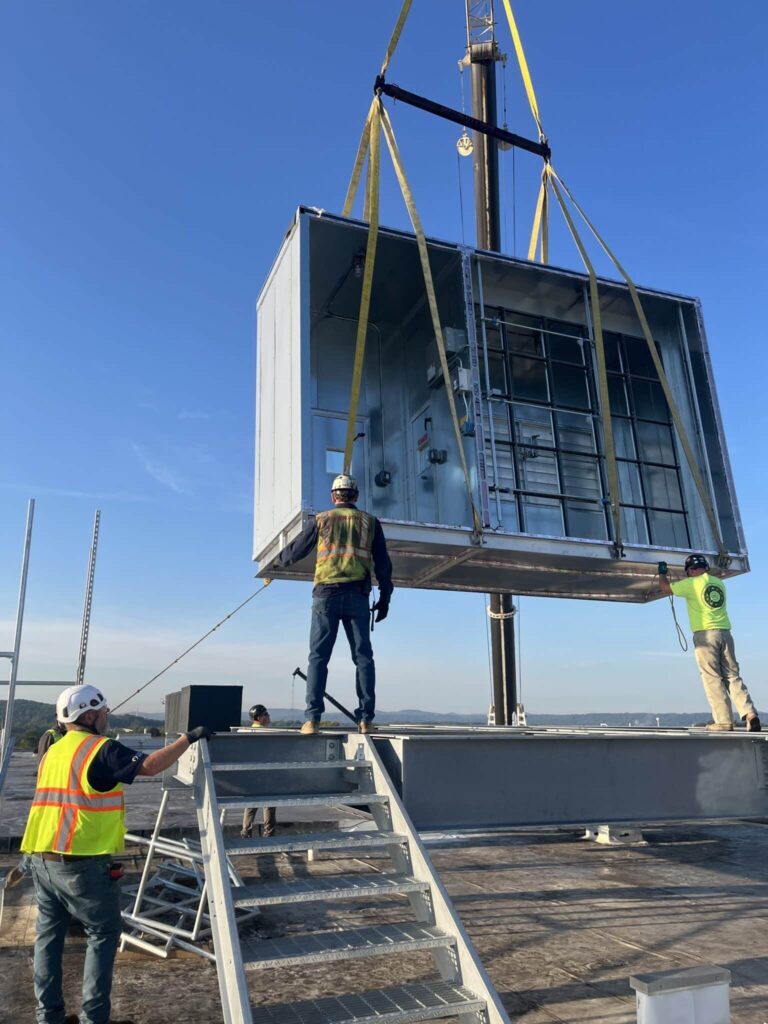 Three construction workers with safety gear standing on a rooftop as they guide a large industrial ventilation unit being lifted by a crane. One worker holds a guide line, ensuring the unit is positioned accurately. The background shows a clear blue sky and distant hills.