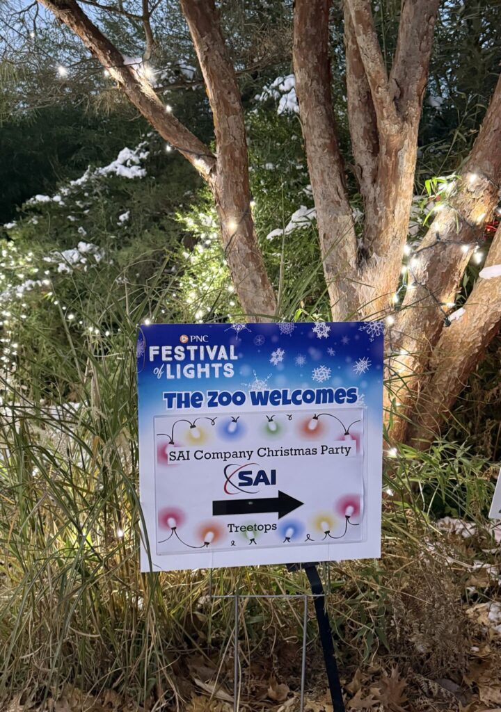 A sign at the Festival of Lights event at the zoo, welcoming attendees to the SAI Company Christmas Party. The sign is illuminated by string lights and surrounded by trees. It has the text 'PNC Festival of Lights' and arrows pointing to 'Treetops' for the party location.
