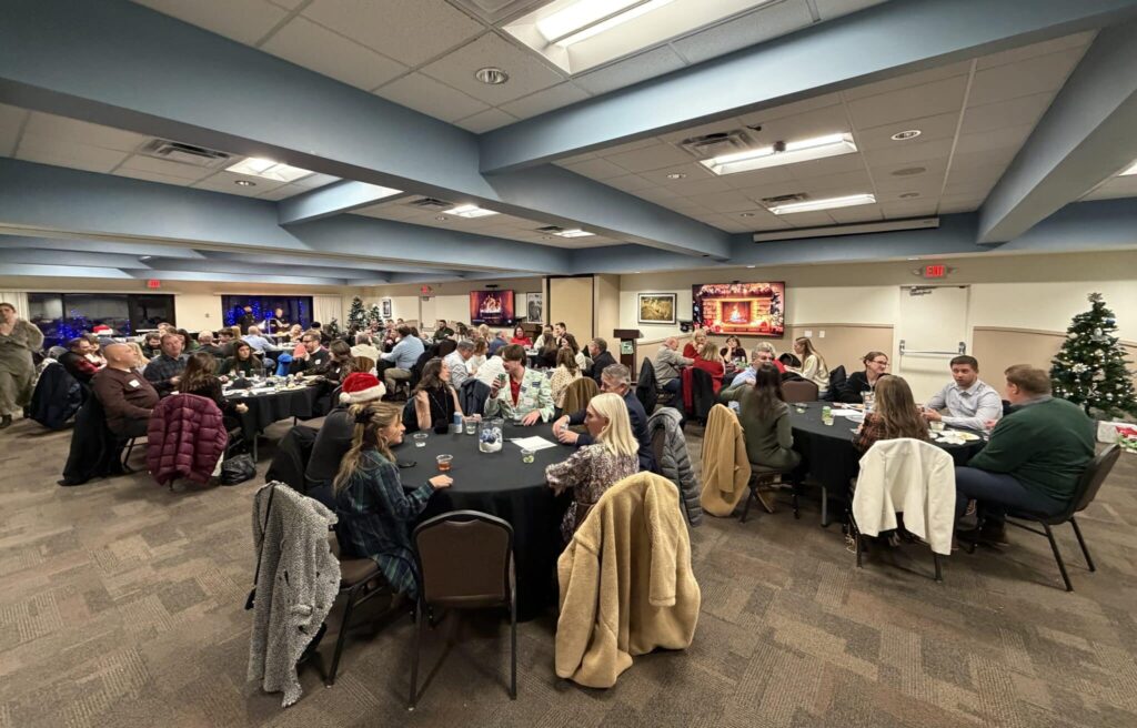 A large group of people seated around round tables in a banquet hall, decorated for a holiday gathering. The tables are covered in black tablecloths, and many attendees are wearing festive attire. There are holiday decorations, including a Christmas tree and a digital fireplace display on the wall, creating a warm, celebratory atmosphere.
