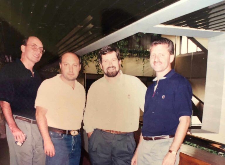 Four men standing together indoors, smiling at the camera. One man is holding a can. They are dressed casually in shirts, with a backdrop of a ceiling and some green plants.