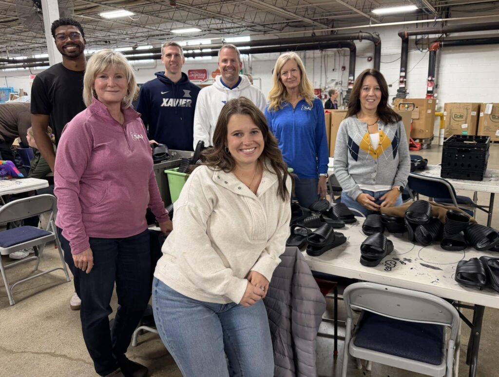 A group of seven people standing and smiling inside a warehouse. They are gathered around tables filled with black footwear. The setting appears industrial with exposed ceiling beams and large boxes in the background. The individuals are casually dressed in sweaters and jackets.