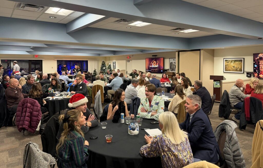 A holiday party in a banquet hall with several round tables filled with people chatting and enjoying drinks. Some guests are wearing festive attire, including Santa hats. The room is decorated with Christmas trees and a digital fireplace image on a large screen. Attendees are engaged in conversation, surrounded by black tablecloths and holiday decorations.
