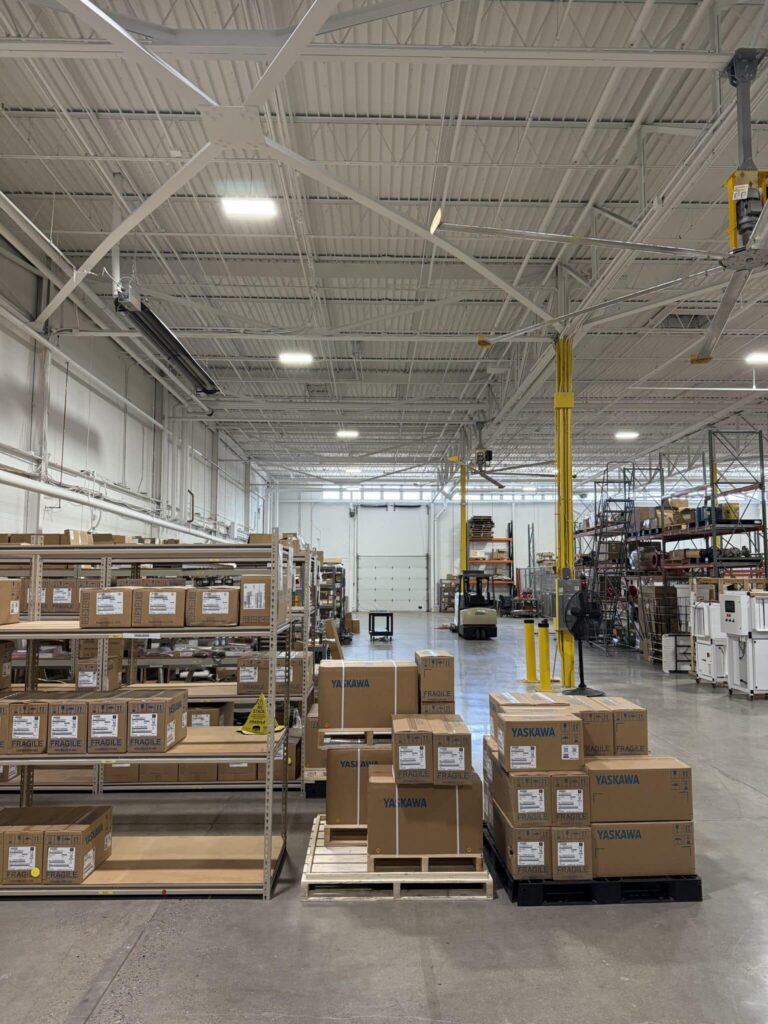 A spacious industrial warehouse with high ceilings and metal beams. The floor is concrete, featuring shelves filled with labeled boxes and multiple pallets of stacked boxes with the brand 'Yaskawa'. In the background, a forklift is visible next to more shelving with various items. Overhead lighting illuminates the facility, providing a well-lit area for storage and operations.