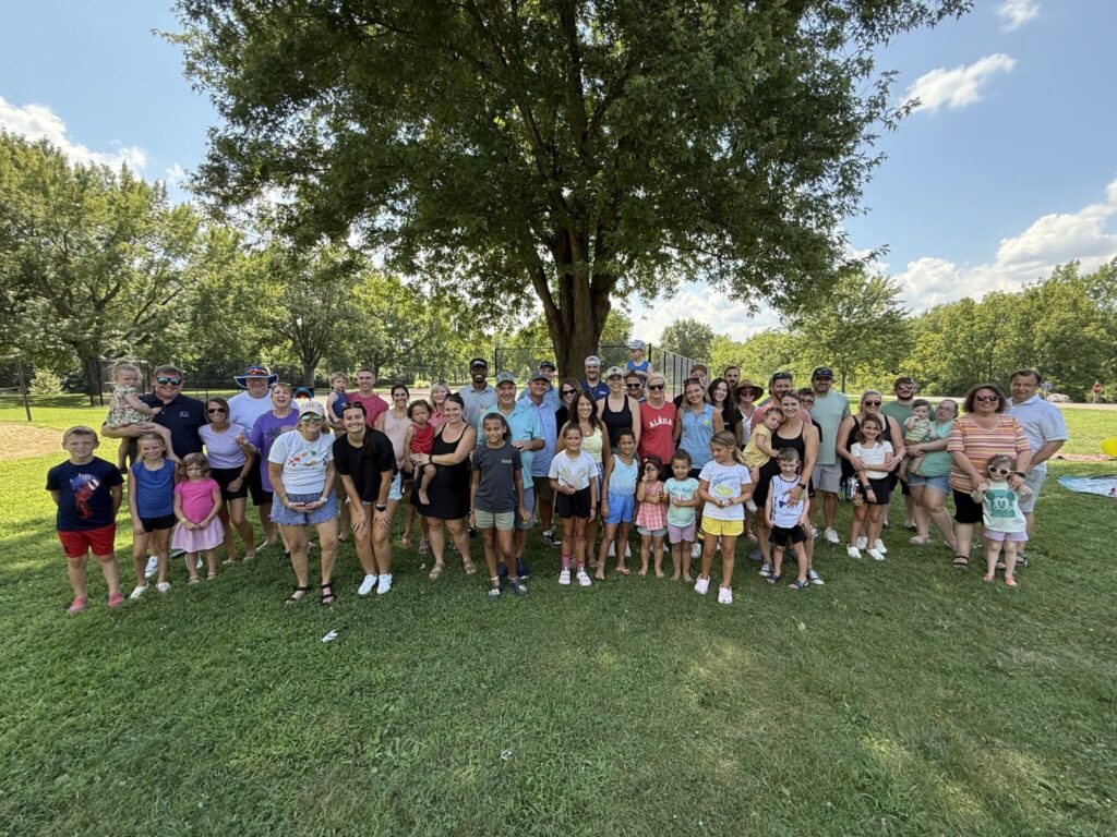 A large group of people, including adults and children, are gathered together in a park under a tree. They are smiling and posing for the photo, with a green lawn and trees in the background under a partly cloudy sky.