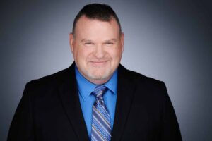 A smiling man wearing a dark suit, blue shirt, and striped tie is positioned against a gradient gray background. The man has short hair and facial stubble, conveying a professional appearance.