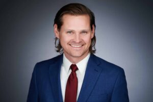 A man with medium-length brown hair, wearing a blue suit jacket, white shirt, and red tie, smiles against a plain gray background.