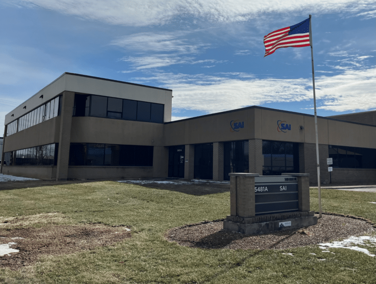 A modern office building with large windows and a beige exterior. The building has a sign displaying 'SAI' and an American flag flying on a pole in the foreground. Snow patches are visible on the grass surrounding the building.