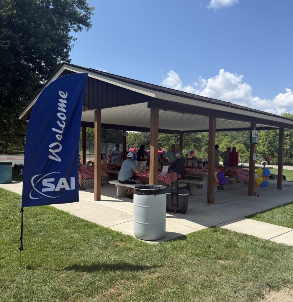 An outdoor picnic shelter with people gathered under it. The shelter has a roof and picnic tables with red and white checkered tablecloths. A blue flag with the word 'Welcome' and the letters 'SAI' is visible on the left. Colorful balloons are tied to the railing, creating a festive atmosphere. The background shows trees and a blue sky with scattered clouds.
