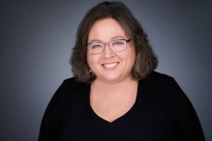A portrait of a woman with shoulder-length hair and glasses, smiling against a neutral gray background. She is wearing a black top and looks directly at the camera.