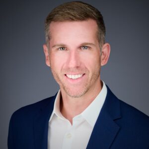 A professional headshot of a man in a blue suit jacket and white shirt, smiling against a gray background.