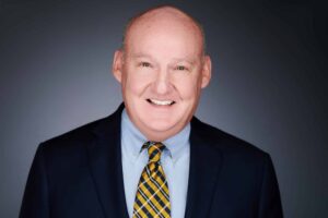A bald man in a business suit and striped tie, smiling at the camera against a dark gradient background.
