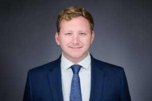 A young man with short hair wearing a navy blue suit and tie, posing against a neutral gray background. He has a slight smile and is looking directly at the camera.