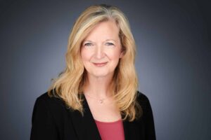 A professional headshot of a woman with long blonde hair wearing a black blazer and a red top, set against a neutral gray background. She is smiling softly and looking directly at the camera.