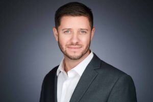 A professional headshot of a man with short dark hair and a beard, wearing a gray suit and white shirt, smiling slightly against a gray background.