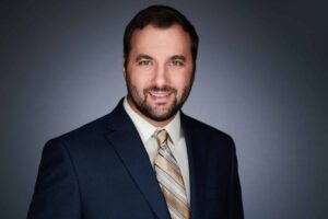 A man with a beard wearing a dark suit and striped tie, smiling against a plain gray background in a professional headshot.