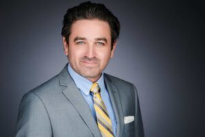 A professional portrait of a man wearing a gray suit, light blue shirt, and a yellow striped tie, with a pocket square visible. He has short dark hair and is smiling softly against a dark gray background.