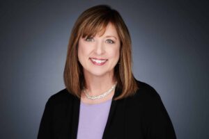 Professional portrait of a woman with shoulder-length brown hair wearing a black blazer, light purple top, and a silver necklace, smiling against a gray background.