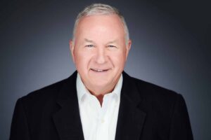 A professional portrait of an older man with short gray hair, wearing a black suit jacket and white shirt, smiling against a dark gradient background.