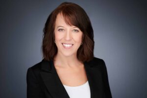 A woman with shoulder-length brown hair, wearing a black blazer and white top, smiles warmly against a gray background in a professional headshot.