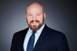 A bald man with a reddish-brown beard and blue eyes, wearing a dark suit and a blue tie with white polka dots, smiling at the camera against a dark gray background.