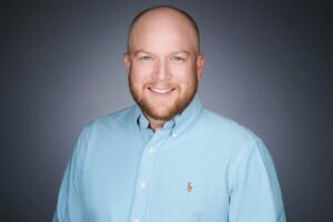 A man with a beard and blue eyes is smiling in a portrait. He is wearing a light blue button-up shirt with a small logo on the left side. The background is a neutral, dark gray color.