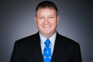 A man wearing a dark suit and light blue shirt with a blue tie featuring white logos, smiling against a plain dark background.