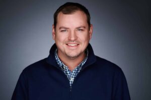 Portrait of a smiling man wearing a dark blue sweater over a blue and white checkered shirt, set against a dark gradient background.