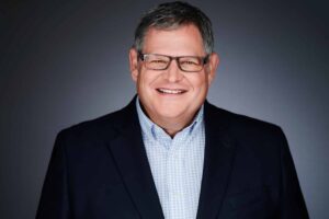 A middle-aged man with glasses smiling, wearing a dark suit jacket over a light blue checkered shirt, against a gradient gray background.