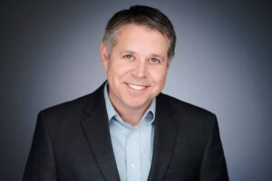 A smiling man with short hair wearing a dark business suit and light blue collared shirt, standing against a neutral gradient background.