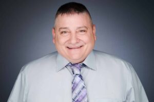 A man with short dark hair smiling, wearing a light grey shirt and a purple plaid tie, against a gradient grey background.