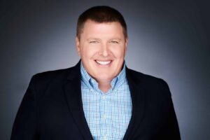 A man with short hair smiling broadly, wearing a dark suit jacket over a blue and white checked shirt, set against a gradient gray background.