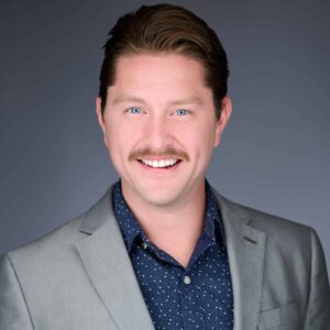 A man with a mustache is smiling broadly. He is wearing a light gray suit jacket over a dark shirt with a subtle pattern. The background is a gradient of dark gray, and the image has a professional headshot feel.