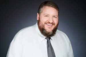 A man with a beard smiling, wearing a white shirt and a striped tie, against a dark background.