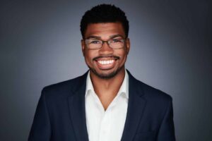 A man with short curly hair and glasses is smiling broadly. He is wearing a dark suit jacket over a white shirt, and the background is a plain gray, giving a professional and friendly appearance.