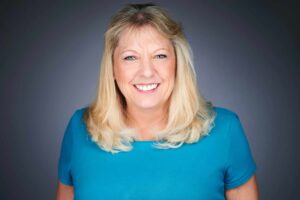 A smiling woman with blond hair wearing a blue shirt, looking directly at the camera against a neutral background.