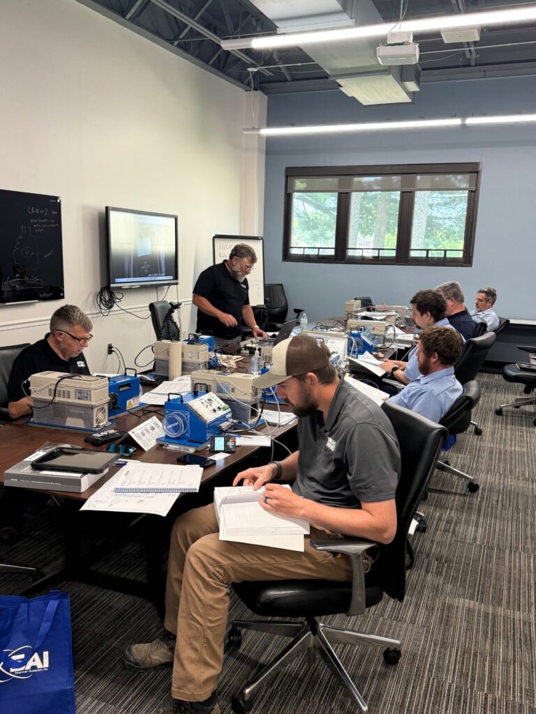 A classroom setting with several individuals seated and engaged in a technical training session. One person is standing and instructing the class, while others are seated at a table with various equipment and manuals. A screen and a whiteboard are mounted on the walls, and there are large windows letting in natural light.