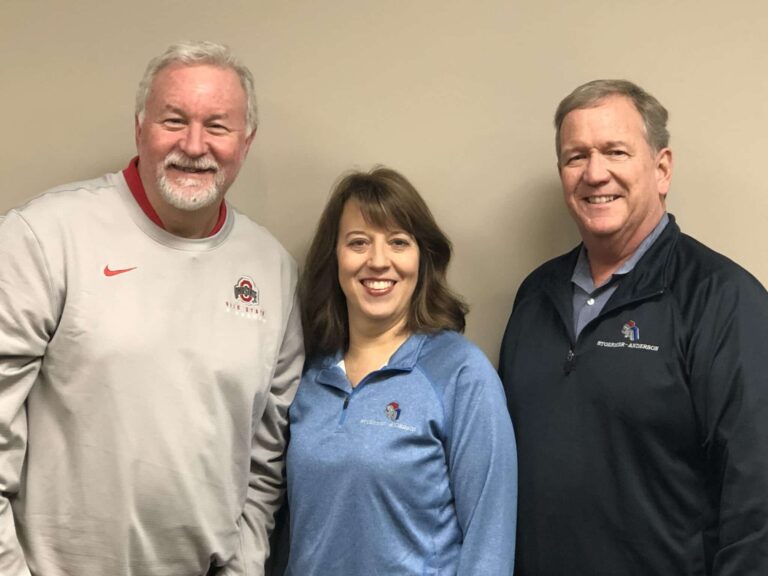 Three people are smiling in a group photo. The person on the left is wearing a light gray long-sleeve shirt with a logo. The person in the middle is a woman in a light blue collared shirt. The person on the right is wearing a dark blue jacket with a logo. They are standing against a plain beige background.