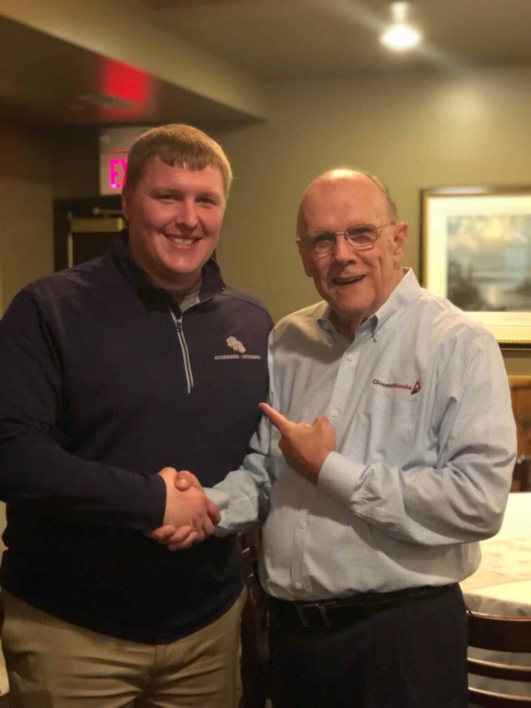 Two men shaking hands and smiling in a warmly lit room. The man on the left is wearing a dark sweater and khaki pants, while the man on the right is in a light blue checkered shirt and glasses, pointing towards the other man. A framed picture hangs on the wall behind them.