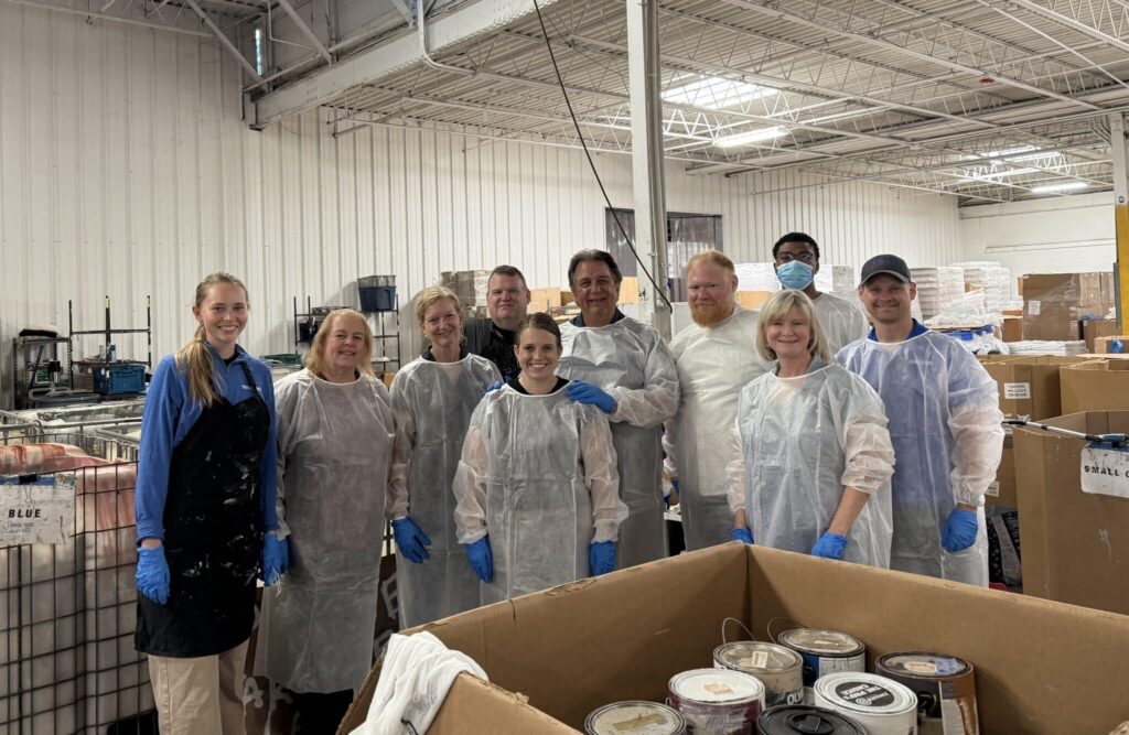A group of ten people wearing protective overalls and gloves standing together in a warehouse environment. The warehouse has high ceilings and industrial shelving with stacked boxes and containers. The individuals are smiling and appear to be volunteers, possibly working with paint cans in front of them.