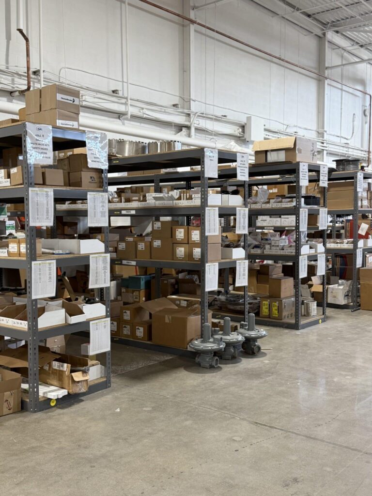 Interior of a warehouse showing multiple industrial shelves filled with various cardboard boxes and parts. The shelves are metallic and organized with labels, and the floor is clean with visible concrete. Overhead pipes and lighting fixtures are seen in the high ceiling.