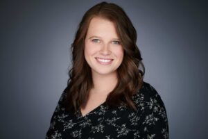 A woman with dark brown wavy hair smiling, wearing a black floral-patterned top, against a plain gray background.