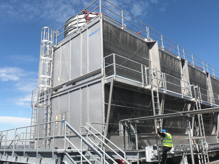 A large industrial cooling tower with a metal structure and ladders, featuring a worker in a safety vest and helmet inspecting the equipment. The sky is clear and blue, highlighting the prominence of the machinery.