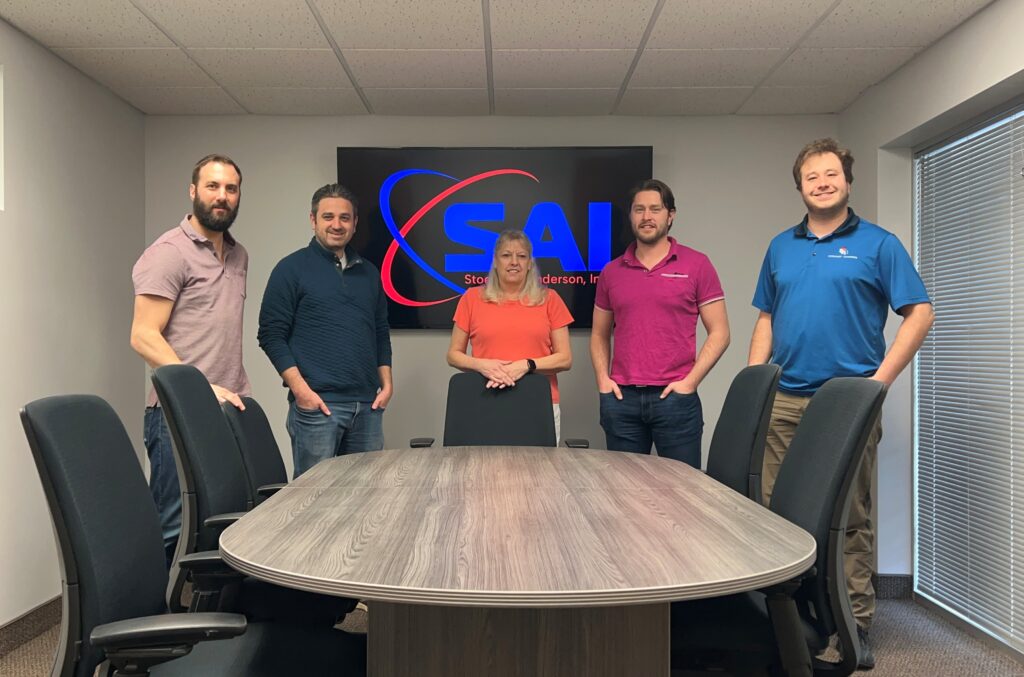 Five people standing in a conference room behind a large oval table. They are in front of a screen displaying the SAI logo. The room has several office chairs and a window with blinds on the right side.