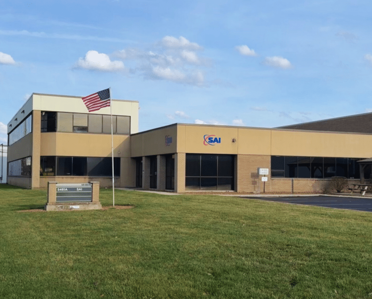 A modern office building with a beige and gray exterior, featuring large windows and the SAI logo on the wall. An American flag is prominently displayed on a pole in front of the building, with a neatly maintained grass lawn and a sign labeled '5481A SAI'. The sky is blue with scattered clouds.