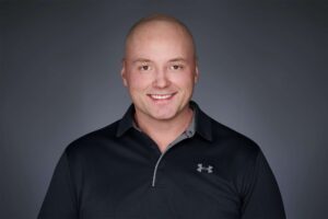A bald man smiling and wearing a black polo shirt with a logo on the chest against a dark gray background.