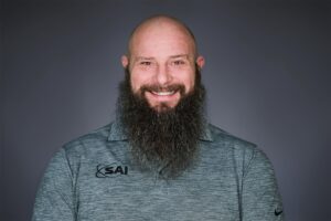 A smiling bald man with a large beard wearing a grey polo shirt with the logo 'SAI' on it, set against a dark grey background.
